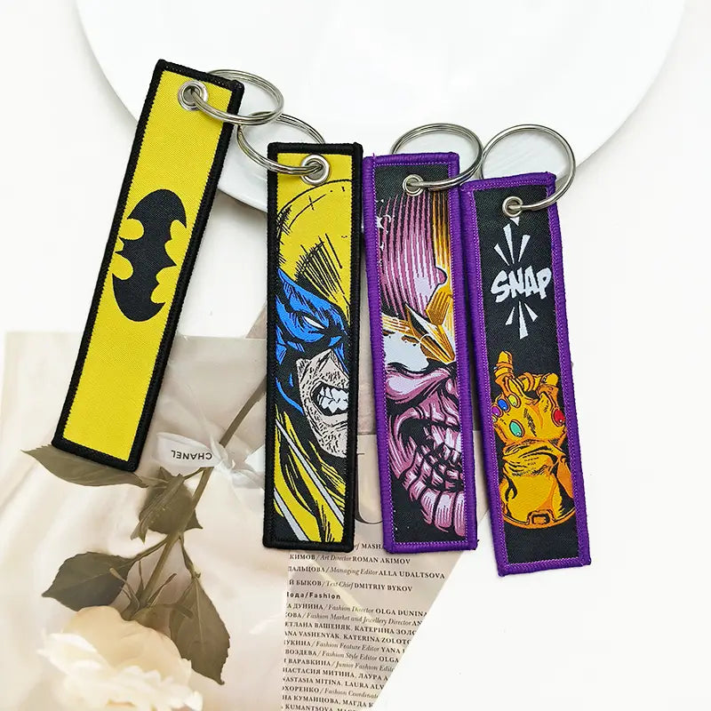 four colorful embroidered superhero keychains featuring batman logo on a yellow background and thanos infinity gauntlet in a superhero embroidered keychain set anime key lanyard