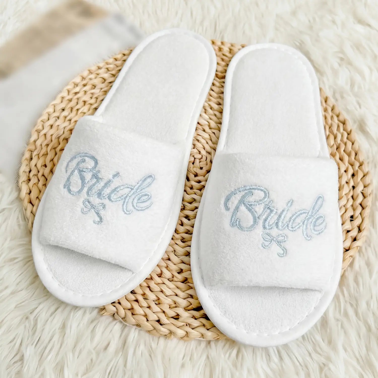 Sandales coulissantes en peluche blanches pour l’équipe de la mariée, avec la mariée brodée en argent, en écriture scintillante, et un petit nœud sous le mot, faisant partie de l’ensemble cadeau pour la mariée de l’équipe.