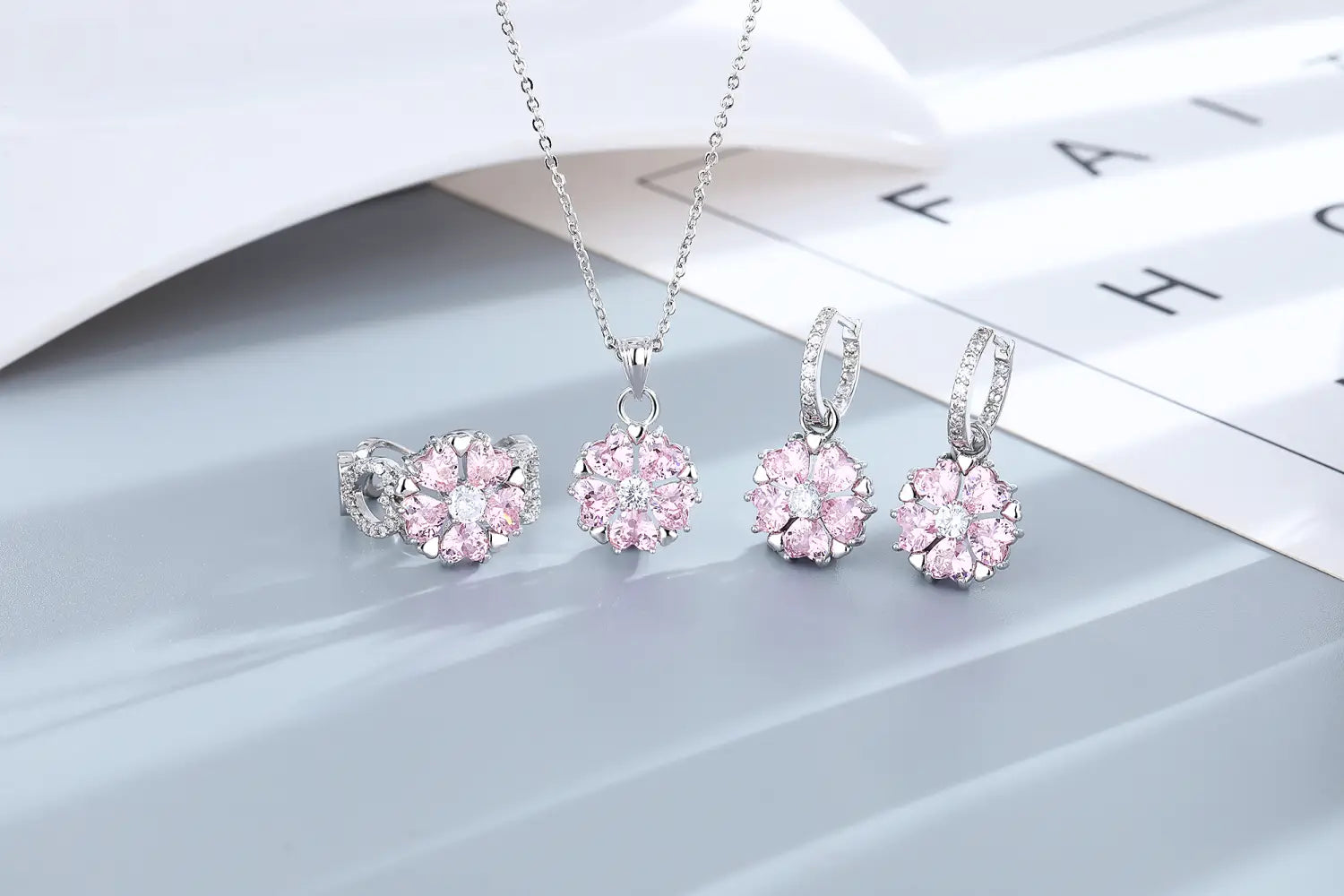 Ein zartes Schmuckset „Silber Pink Kirschblüte“ mit Anhänger, Ohrringen und Ring, besetzt mit funkelnden pinkfarbenen Edelsteinen, die in floralen Designs angeordnet sind.