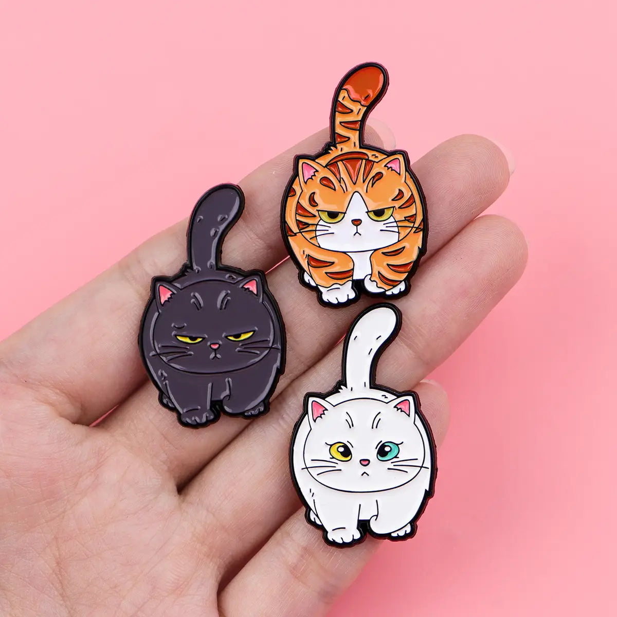 Three cartoon cat enamel lapel pins in black orange tabby and white with expressive eyes and a glossy finish.