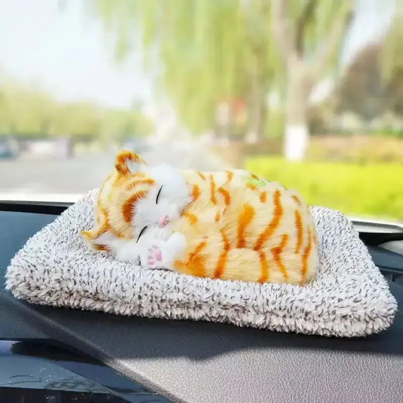 A plush orange and white tabby simulated sleeping cat toy with sound resting on a textured gray and white fuzzy car dashboard mat.