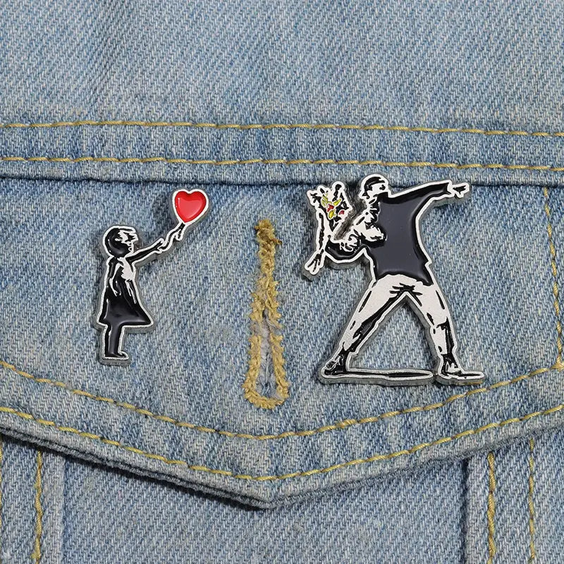 Deux épingles émaillées présentant des motifs d’art de rue emblématiques de Banksy : une fillette tenant un ballon rouge et une silhouette dansant sur un tissu en denim, dans la collection de broches à épingles émaillées « street art » de Banksy.