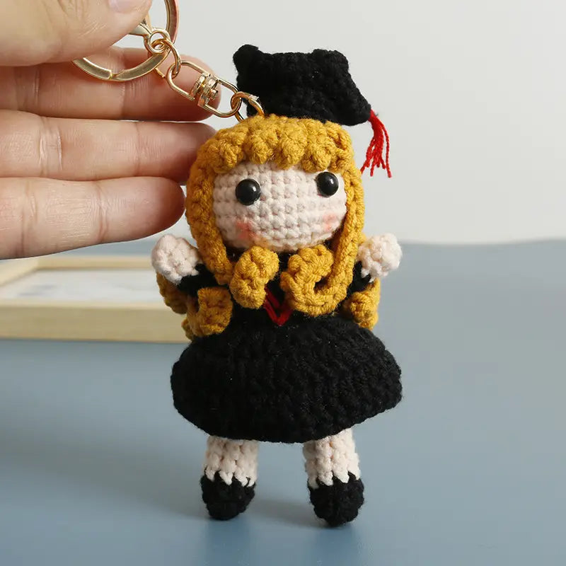 Crochet graduation doll keychain featuring a miniature figure with golden yarn hair wearing a black crochet gown and cap with a white face and rosy cheeks attached to a gold-toned metal ring.