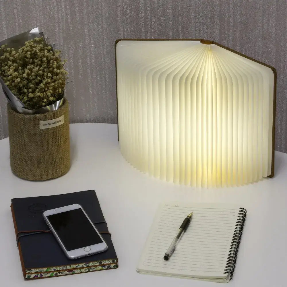 Luz LED plegable con forma de libro con apariencia de libro abierto, con páginas estilo acordeón que emiten un brillo blanco cálido, mostrada desde un ángulo frontal.