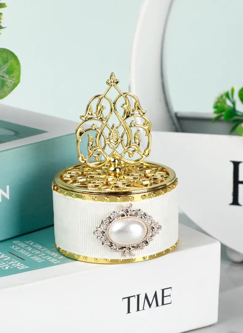 Une boîte à bonbons de mariage dorée blanche cannelée avec bordure dorée, et un motif de perle décorative au centre, surmonté d’un couvercle en filigrane doré orné, utilisée comme contenant de perle-bonbon décorative.