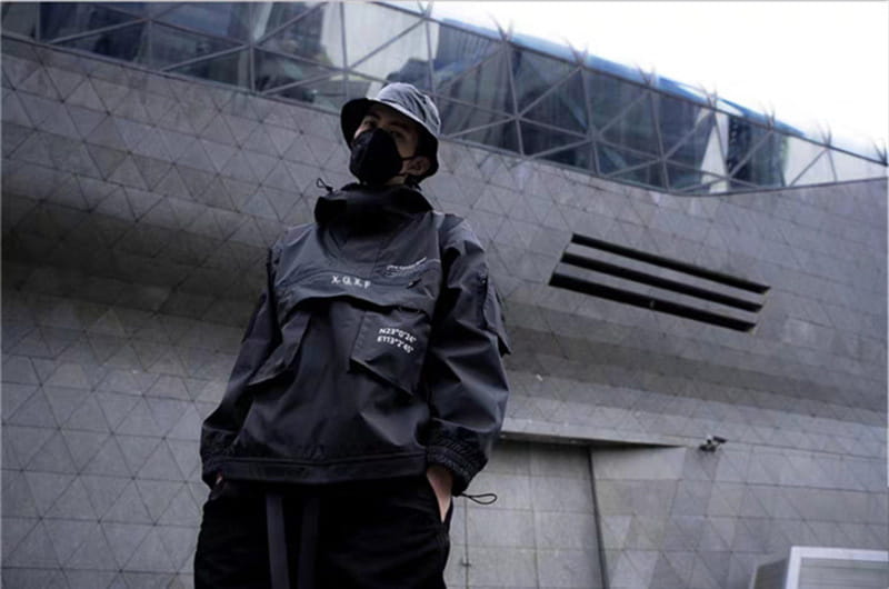 Veste imperméable à capuche tactique techwear noire, avec marquage blanc discret et une capuche portée au-dessus d’un pantalon foncé.
