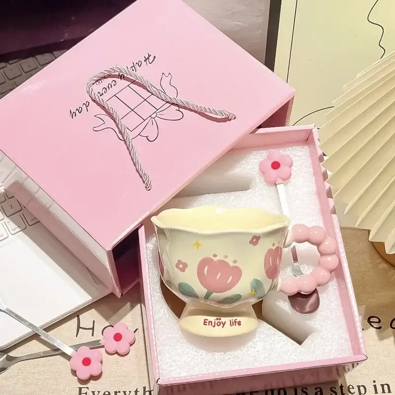 Une tasse tulipe en céramique jaune pastel, avec des décors floraux roses et une anse assortie en perles roses, placée dans un insert en mousse blanche douce à l’intérieur d’une boîte cadeau rose ouverte.