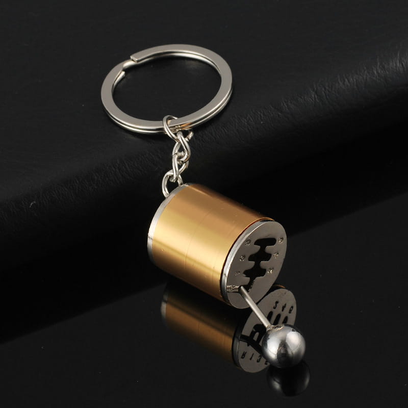 A metallic manual gear shift car keychain with a gold-colored cylindrical body, silver accents, and an engraved gear pattern on its side.