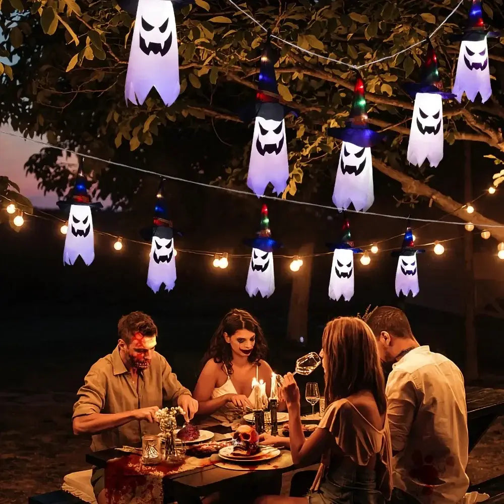 Décoration d’Halloween suspendue en forme de fantôme à DEL, comprenant des lanternes-fantômes gonflables blanches avec des traits du visage noirs et des lumières à DEL multicolores suspendues aux branches d’arbres.