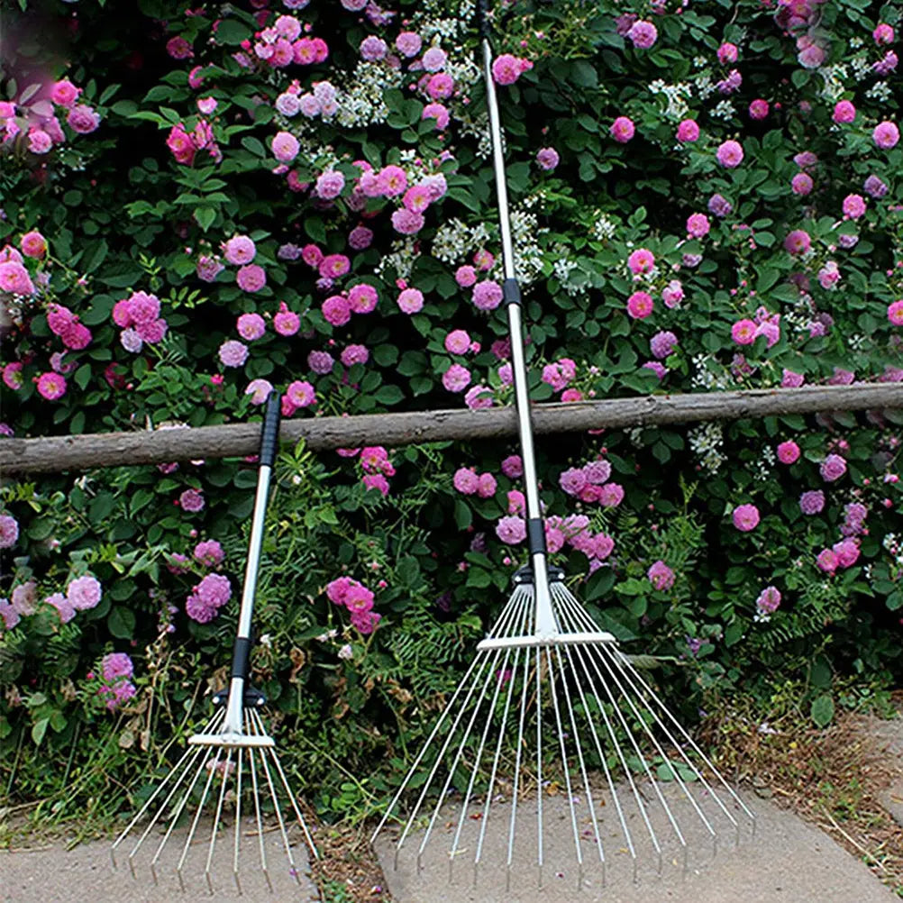 Two silver metal garden leaf rakes with black telescopic handle design, one shorter and one taller, leaning against a wooden fence in front of pink roses.