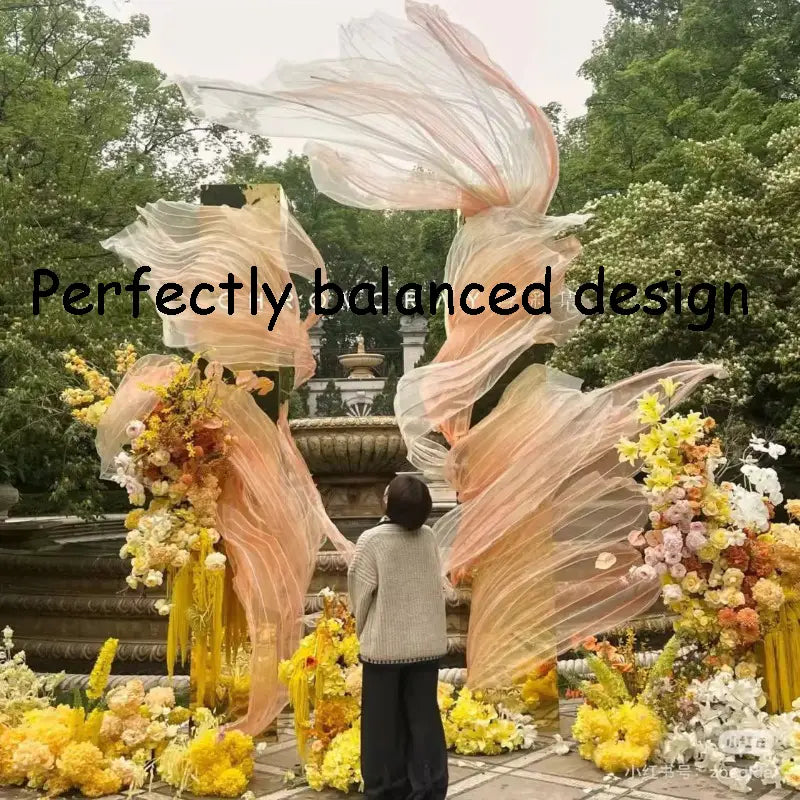 un telón decorativo de guirnalda floral gigante de gasa que presenta un fondo decorativo con forma de flor, con una tela semitransparente fluida de tonos melocotón y arreglos florales vibrantes en amarillo y rosa suave en cascada