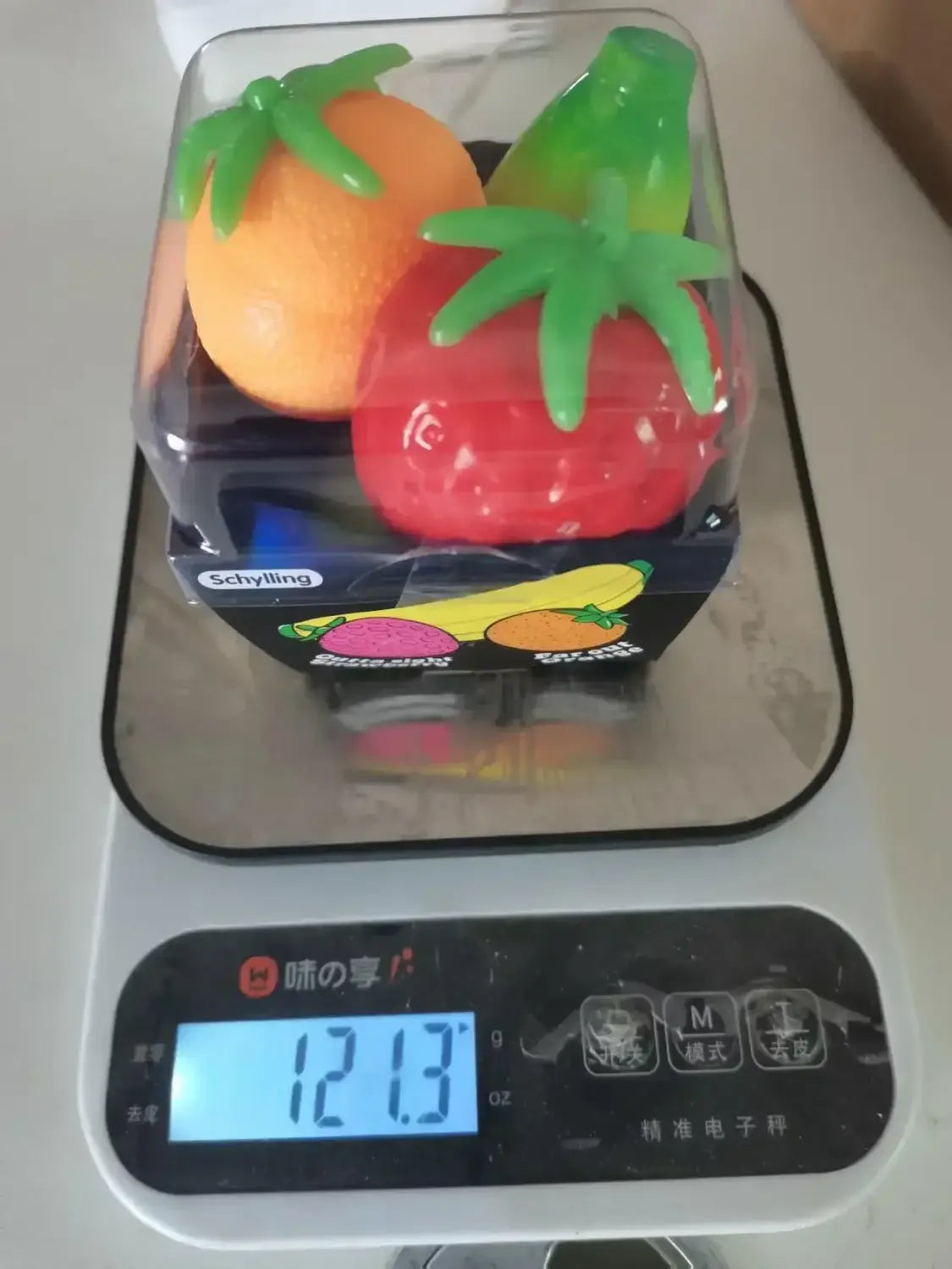 Ein transparenter Kunststoffbehälter mit drei farbenfrohen, quetschbaren Obstformen: eine Orange mit grünem Stiel, eine rote Tomate mit grünem, blättrigem Oberteil und eine teilweise sichtbare gelbe Banane, aus dem Erdbeere-Orange-Banane-Stressabbau-Set.