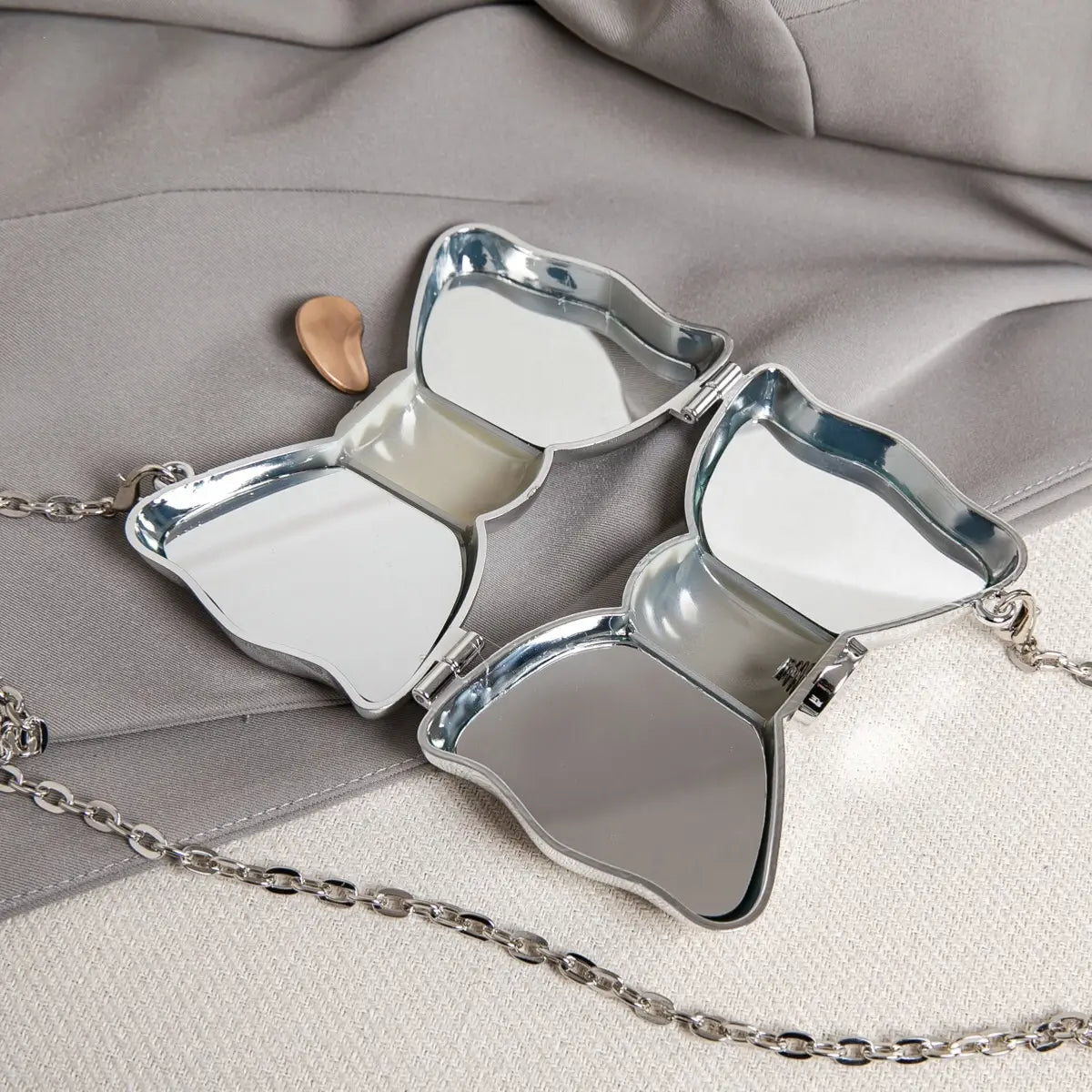 pochette miroir en forme de papillon argentée, finition métallique réfléchissante et anse à chaîne, présentée comme un sac à bandoulière pochette en nœud en métal doré.