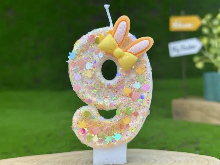 A glittery number nine birthday candle made of wax featuring a yellow bow and orange bunny ears for birthday celebrations.