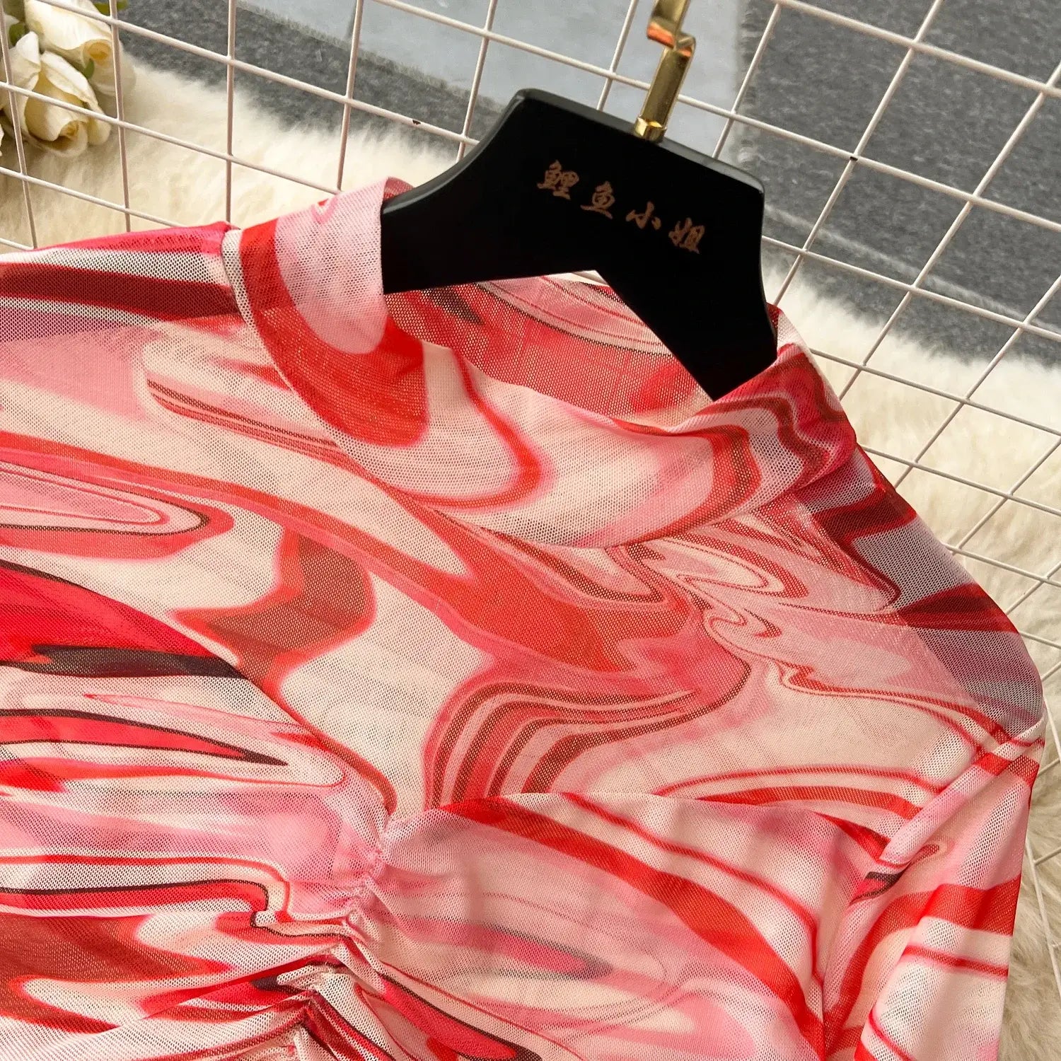 A flowing sheer blouse with a vibrant red and pink marbled pattern hangs on a black hanger as part of a two piece mesh skirt set marble print with elastic closure type.