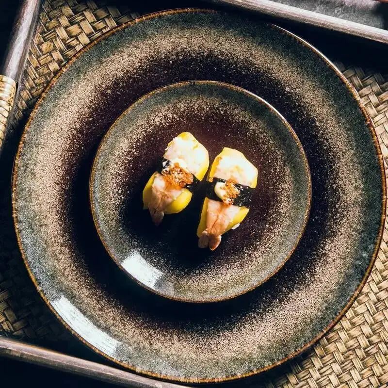 Two pieces of nigiri sushi with yellow fish, pink shrimp, and black seaweed are artfully arranged on a rustic dark brown Japanese ceramic dinner plate.