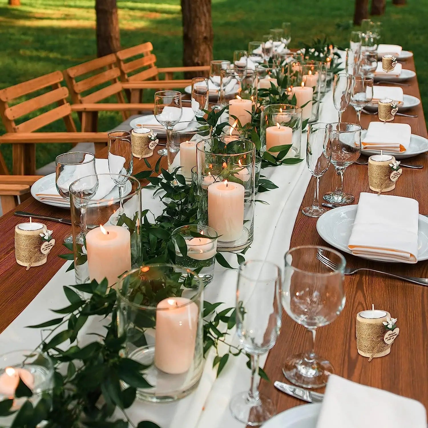 Des bougeoirs en bois pour petites bougies chauffe-plat, en tant que cadeaux d’invités de mariage, présentés comme composition centrale d’une longue table avec de hautes bougies couleur pêche dans des supports en verre transparent et une végétation verte fraîche.
