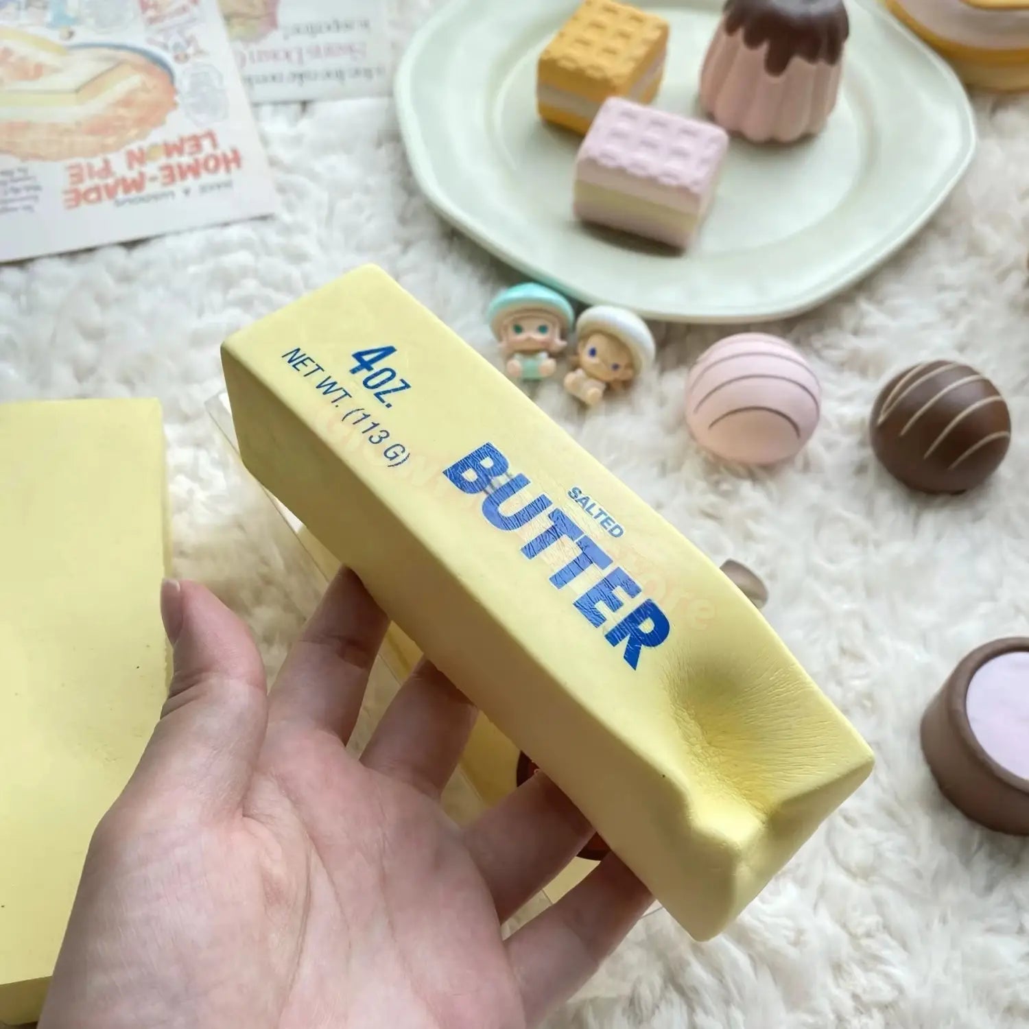 Un jouet antistress en bâton de beurre jaune à montée lente, avec lettrage bleu, tenu dans la main et comprenant la fonction de montée lente, en coffret de 12 pièces.