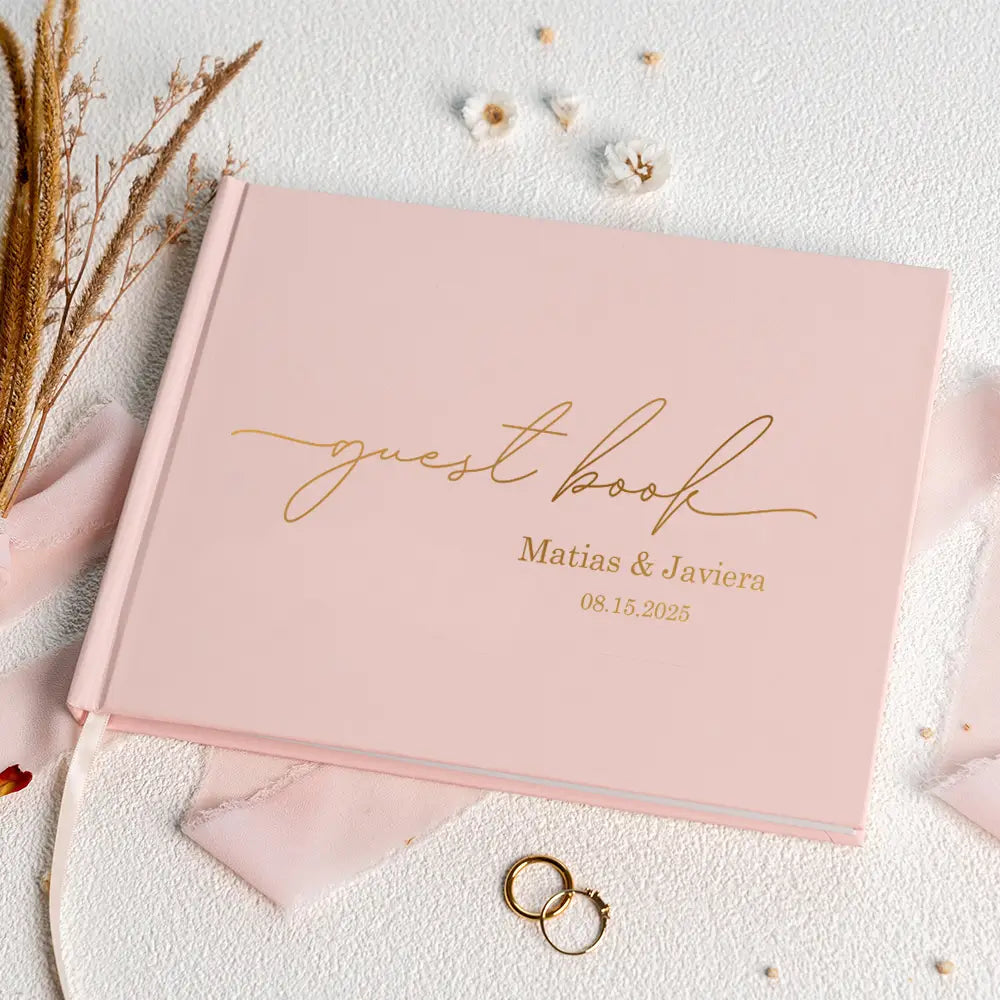 A soft pink personalized wedding guest book with elegant gold cursive lettering, featuring customized names and date, accompanied by a pen.