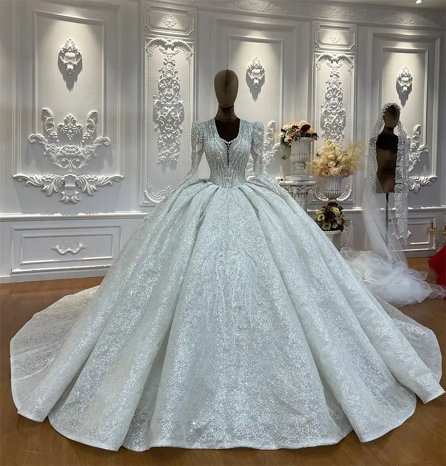 Robe de mariée sur mesure à manches longues en dentelle et jupe en forme de cloche, argenté scintillant, avec jupe ample présentée sur un mannequin dans une pièce blanche ornée.