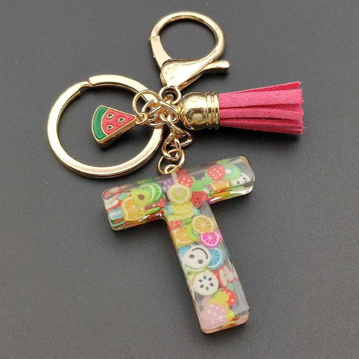 A clear transparent resin-filled letter t keychain with colorful fruit and smiley face charms inside featuring a gold-tone metal ring clasp and pink suede tassel from the fruit letter resin keychain initial alphabet key ring collection.