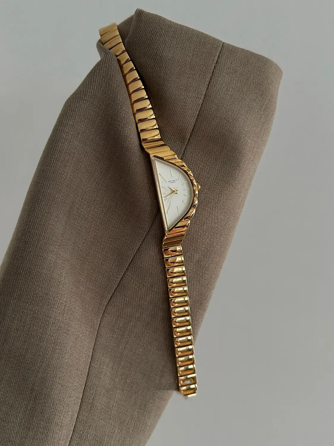 Gold-toned geometric triangle quartz watch with a stainless steel segmented band resting on taupe fabric.