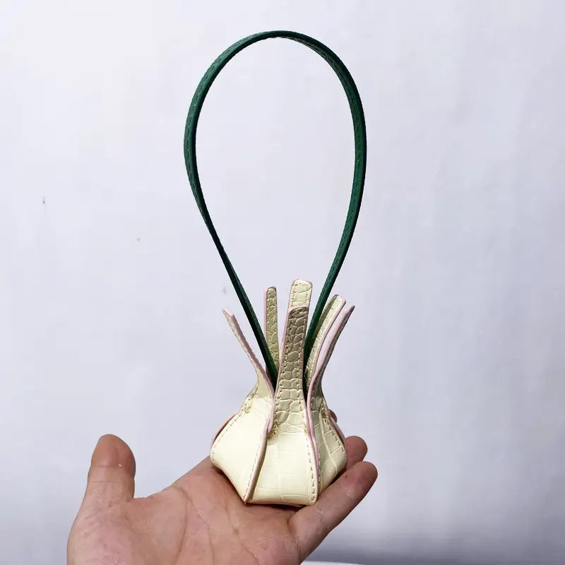 Une pochette en cuir végétal texturé façon crocodile, couleur crème, avec une poignée vert foncé tenue avec délicatesse dans la main, adaptée comme ornement de sac à main de créateur.