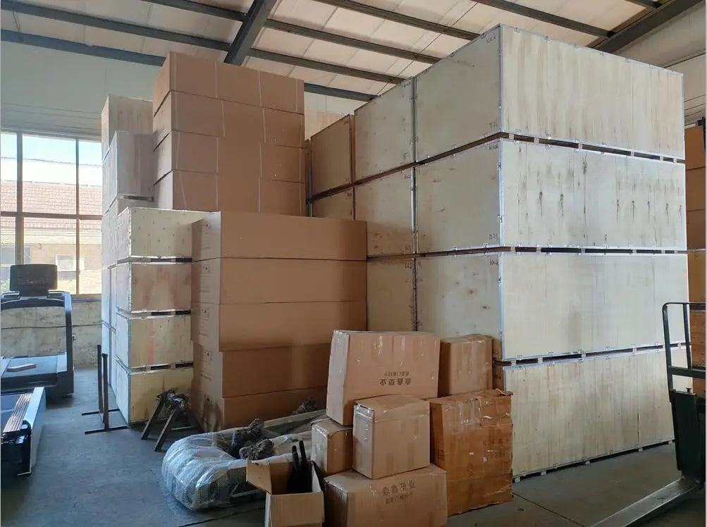 Stacks of light brown cardboard boxes and larger unfinished wooden crates fill the warehouse space where a commercial 2-tier dumbbell rack made of durable materials is stored.