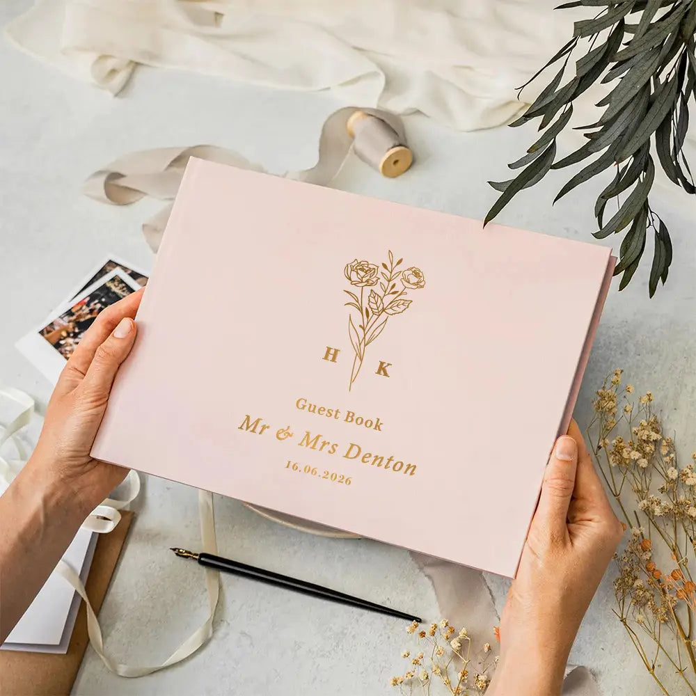 A soft pink matte finish personalized wedding guest book with gold foil floral detailing and initials hk above mr and mrs denton dated 16.06.2020 included with a pen.
