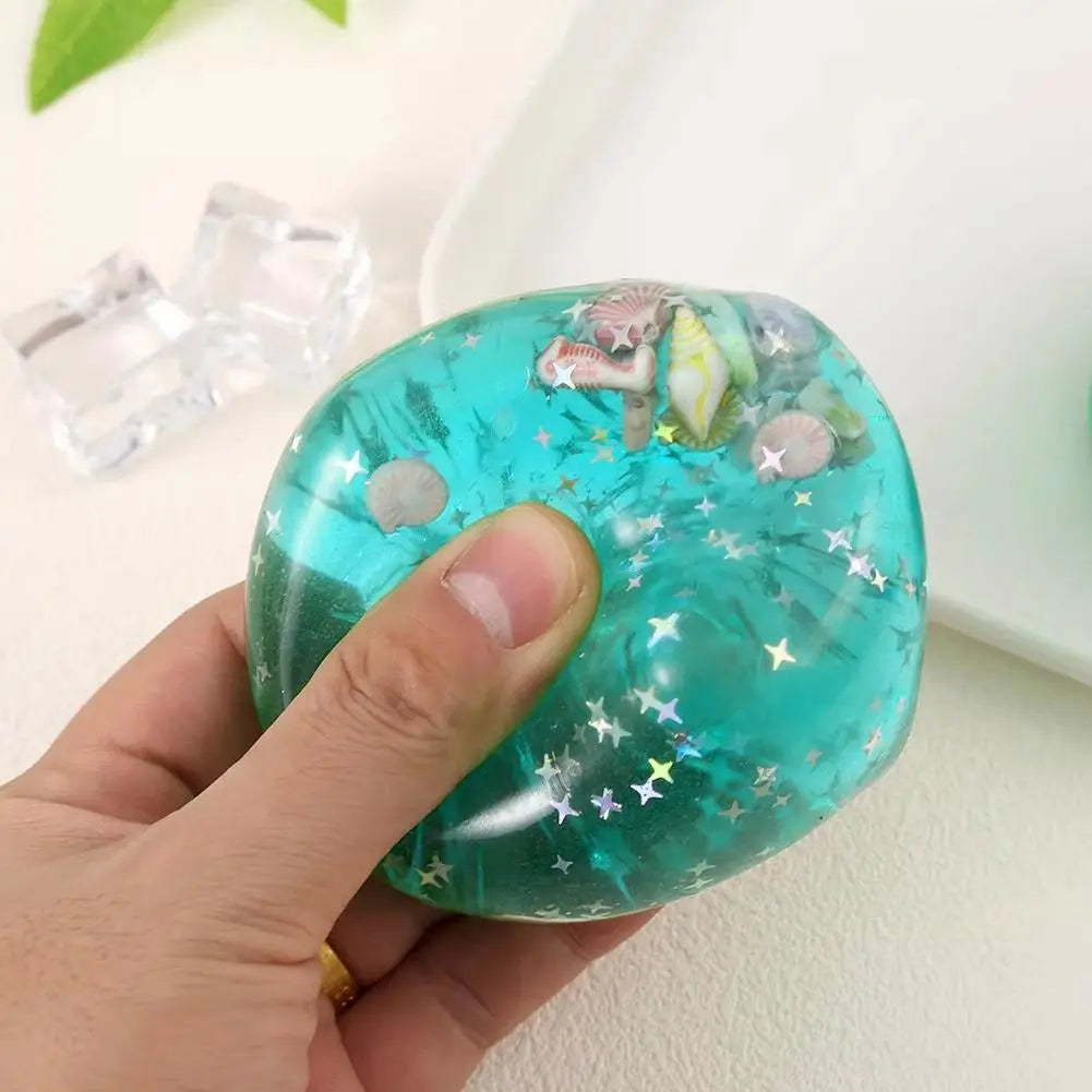Un cube sensoriel à effet anti-stress en océan turquoise translucide, à texture souple, rempli d’étoiles argentées scintillantes et de charmes de coquillages intégrés, conçu comme un cadeau attentionné.