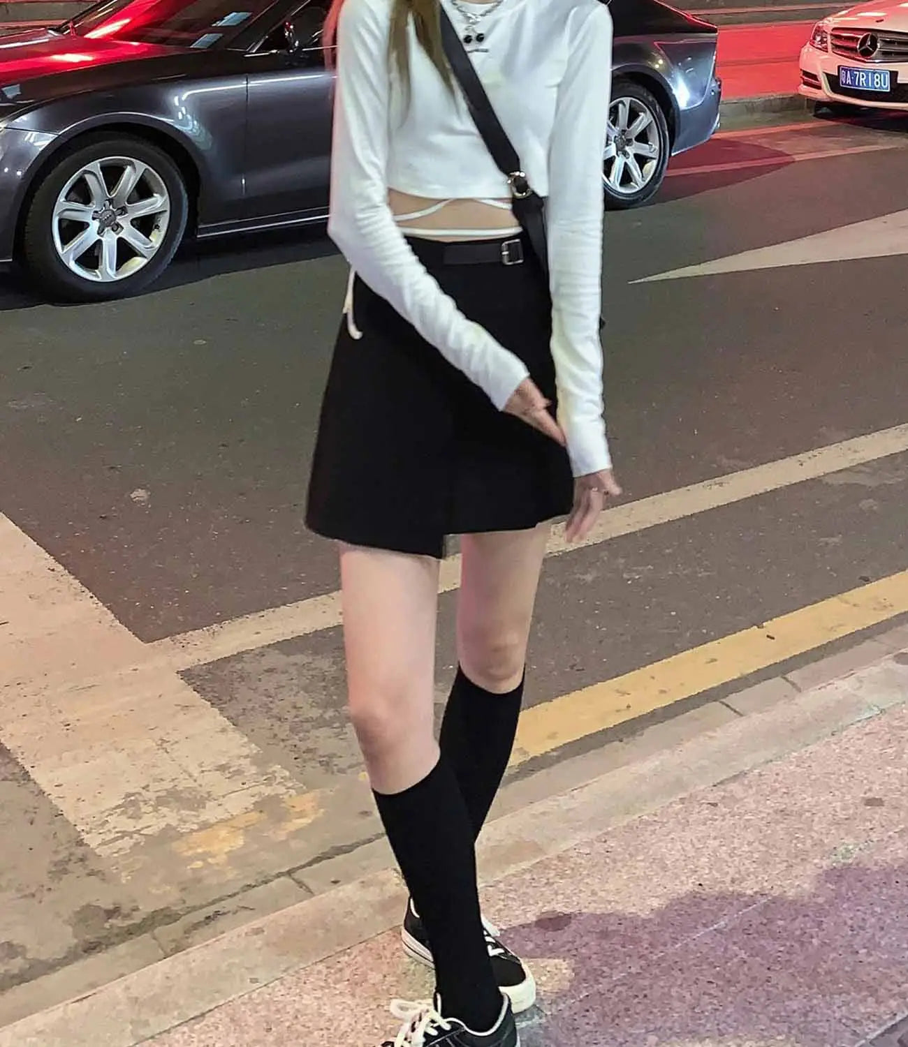 Eine Frau mit einem schwarzen plissierten Minirock, kombiniert mit einem weißen Langarm-Crisscross-Crop-Top und schwarzen Kniestrümpfen
