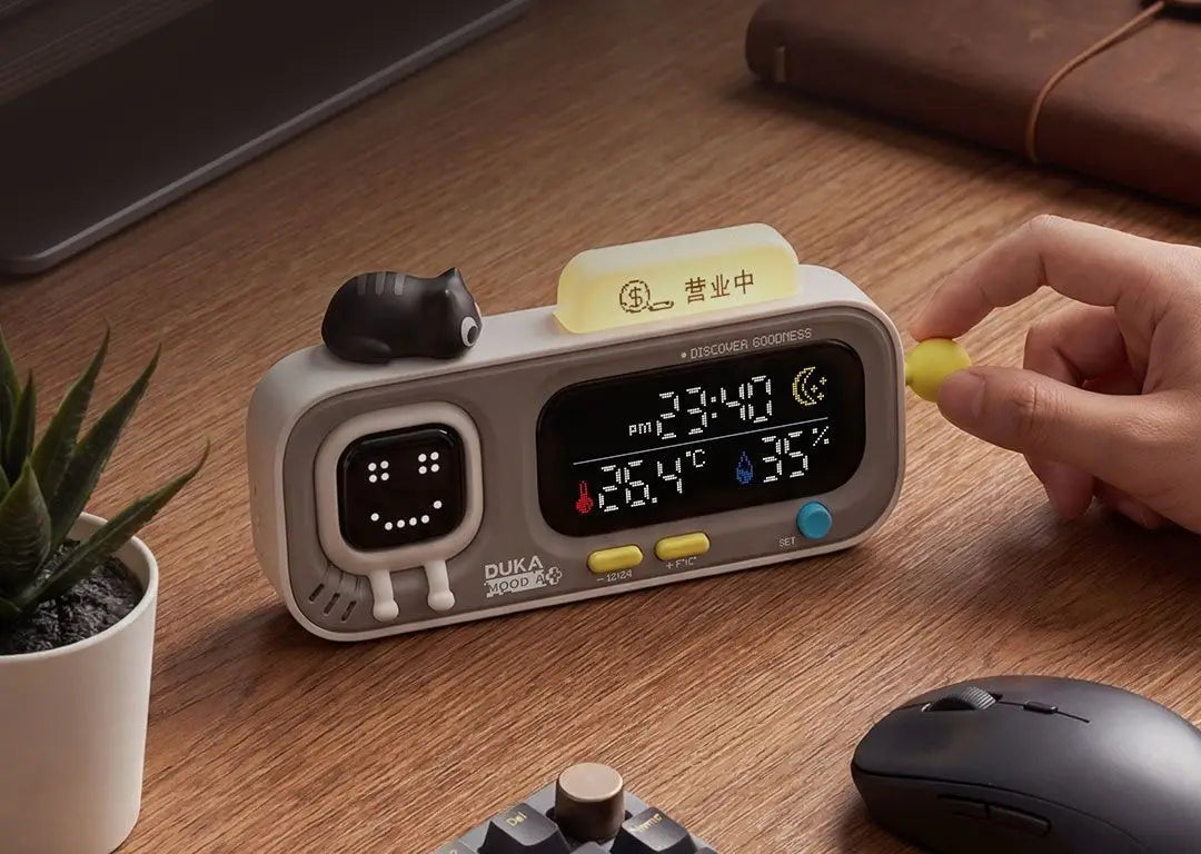 Gray duka mooda smart temperature digital clock with a smiling face display showing temperature and humidity readings and a small black cat figurine on top.