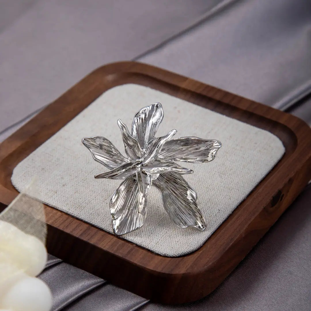A delicate silver flower-shaped brooch with intricately textured petals displayed on a wooden tray alongside an adjustable double layer flower ring.