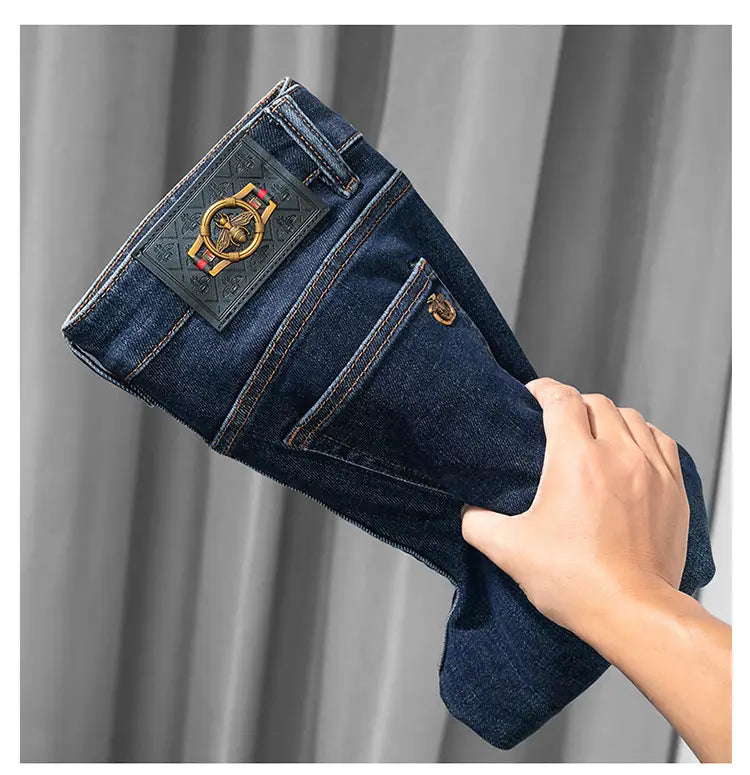 Slim fit dark blue denim jeans for men feature a prominent black leather patch with an ornate gold-colored bee emblem and red thread accents on the back.