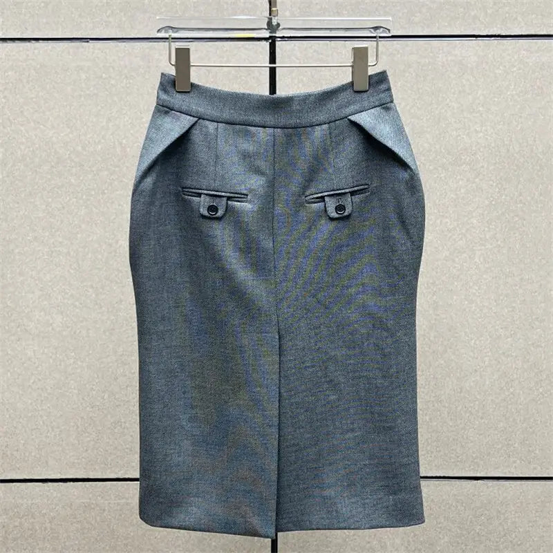 Jupe crayon grise taille haute en laine, sur mesure, de longueur genou, deux poches à rabat frontales fermées par des boutons, et un discret détail de couture sur le côté.