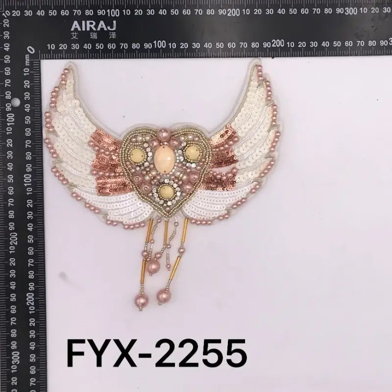 Pièce d’ornement pour sac, broche en forme d’aile d’ange en perles, présentant un appliqué cœur ailé brodé de manière élaborée dans des tons crème et or rose, avec des détails en perles.