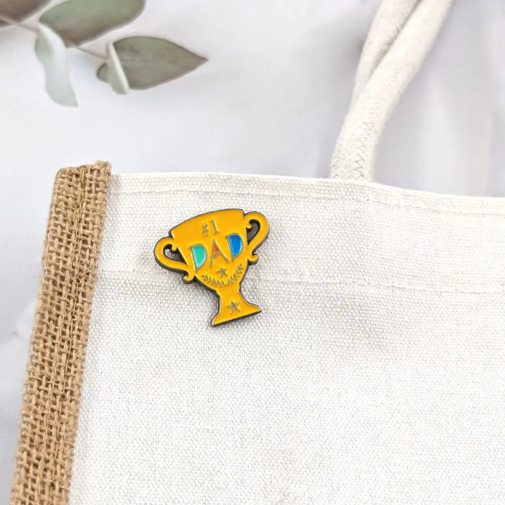 Broche en émail jaune éclatant en forme de trophée avec « number one dad » inscrit en lettres bleues et dorées, rehaussé de petites étoiles conçues comme une broche dorée.