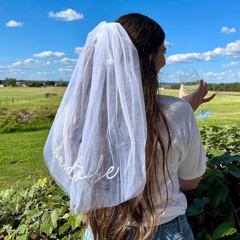 繊細なホワイトのチュール製ベールに、「Bride to be（花嫁になる方）」をエレガントな筆記体で刺繍した、花嫁になる方パーティーセットのベールサッシュおよびヘッドバンドの一部。