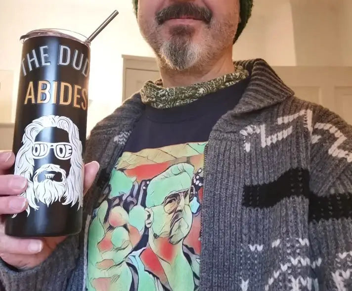 Gourde isotherme noire pour tumbler, avec lettrage doré et un visuel blanc d’un homme barbu portant des lunettes de soleil, avec le texte « the dud abides » affiché à côté d’un pull-over cardigan vintage à fermeture éclair géométrique pour homme.