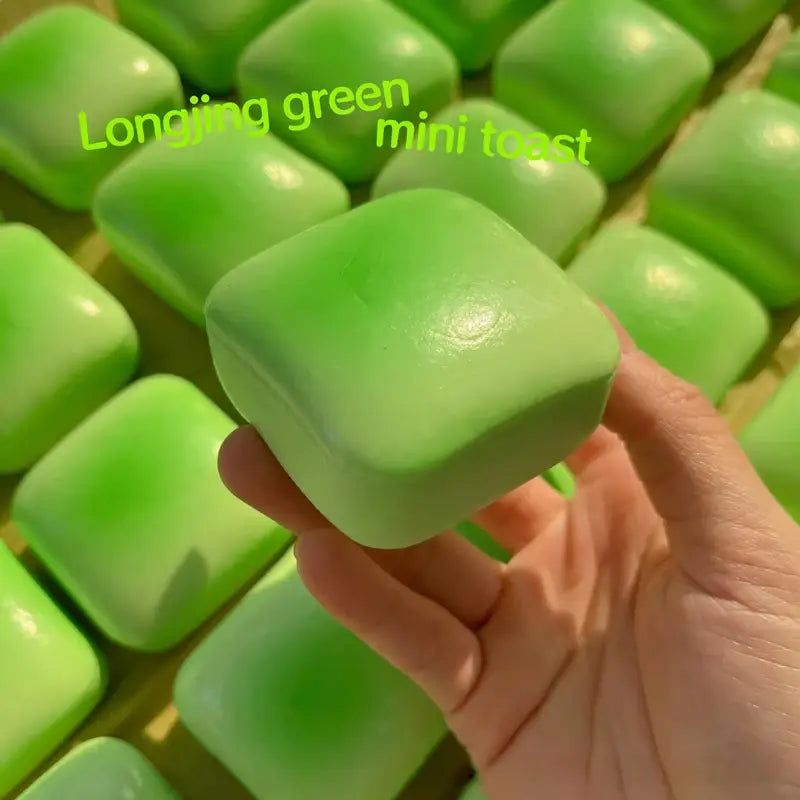 A hand holds a vibrant glossy green longjing green mini toast slow rising squishy toy with smooth rounded square edges.