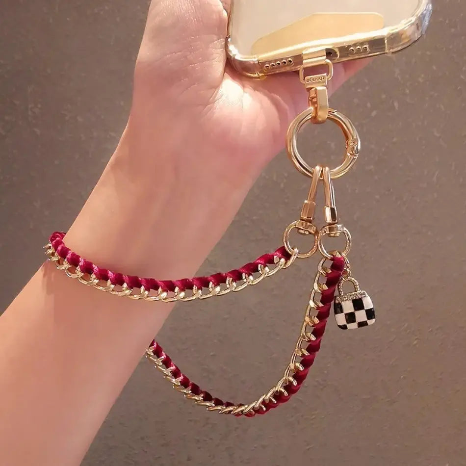 Ein goldfarbener Handy-Handgelenkriemen mit geflochtenem rotem Kord, Kettengliedern und kleinem schwarz-weiß kariertem Schloss-Anhänger zur Verwendung für Anti-Lost-Handylanyard.