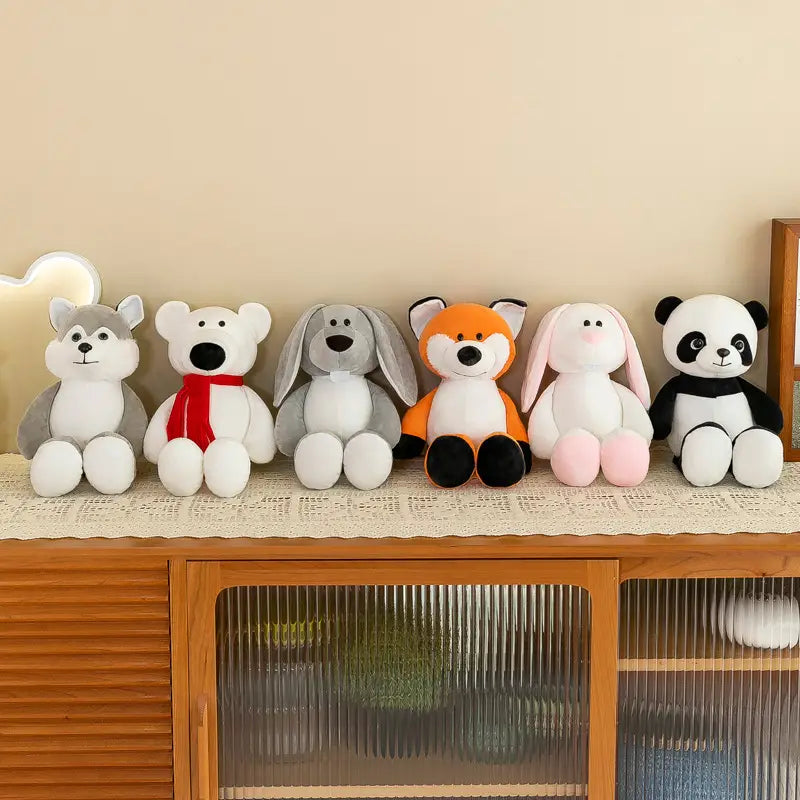 Un rang de six peluches d’animaux de la jungle, représentant notamment un husky gris et blanc, un ours polaire blanc avec une écharpe rouge, un chien gris aux longues oreilles tombantes, et un renard orange aux pattes noires, fabriquées dans une matière souple.