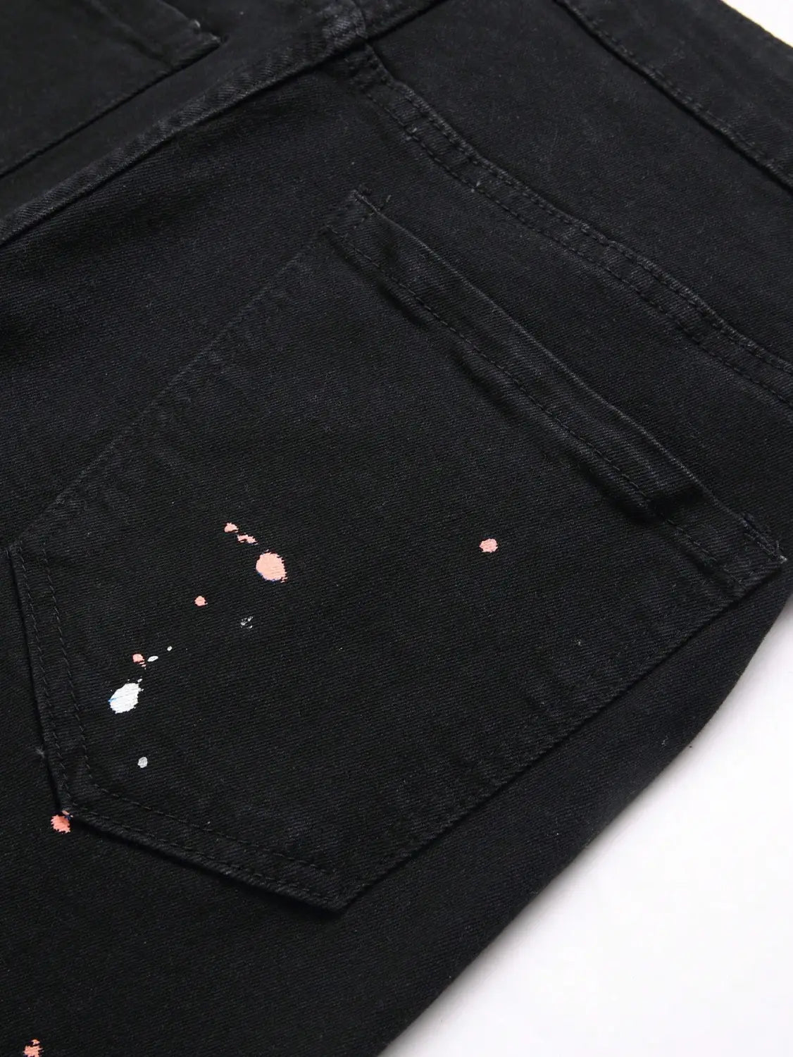 Jeans denim skinny extensibles déchirés Y2K pour hommes en noir, présentant une poche arrière délibérément vieillie, avec des accents d’éclaboussures de peinture rose et blanche.