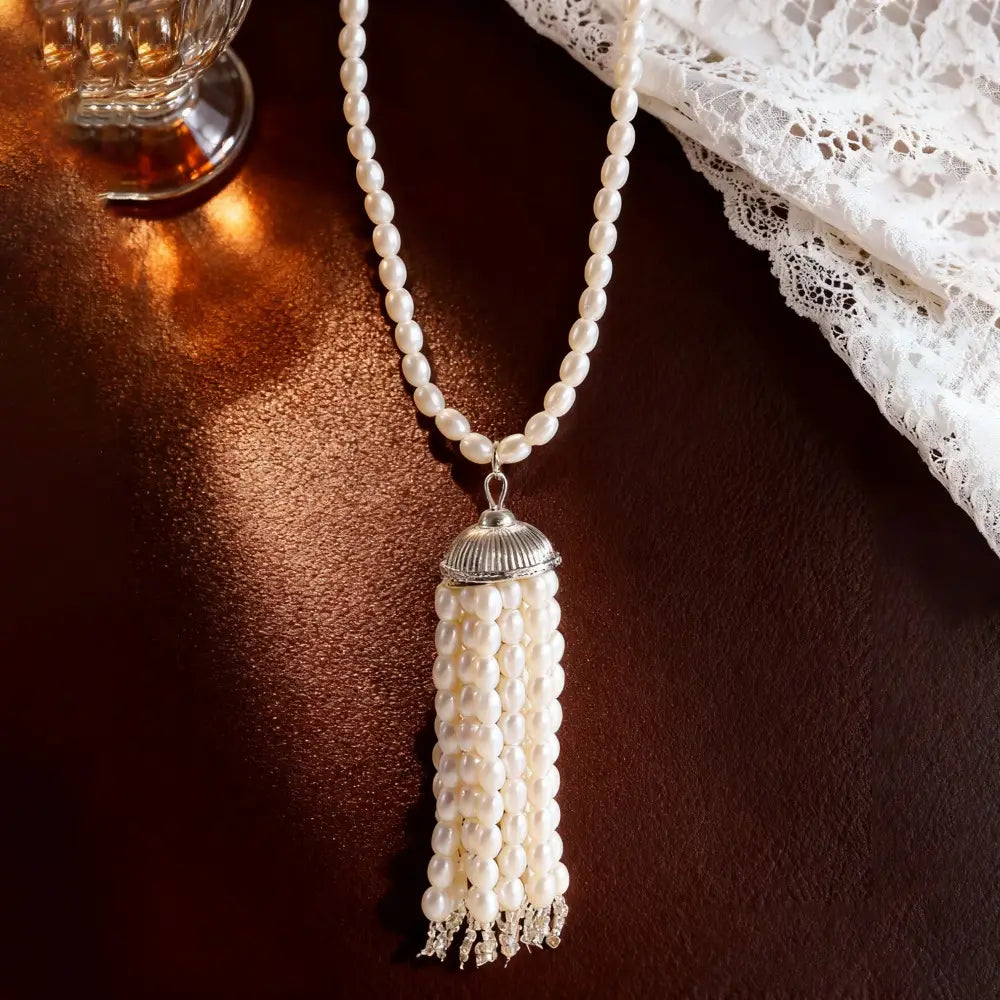 Un délicat collier-ras-de-cou à pendentif pompon en perles blanches, avec une coupelle cannelée à ton argenté et de minuscules accents en cristal, conçu comme une longue chaîne de pull-over.