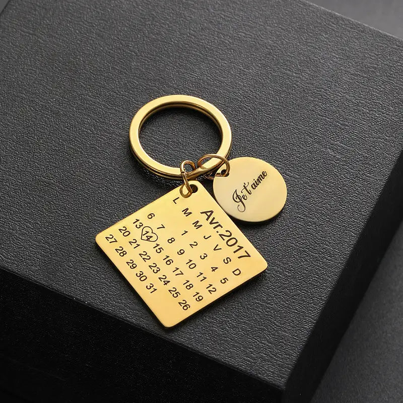Gold-colored metal personalized calendar keychain featuring a square charm engraved with avr.2017 and dates alongside a round disc inscribed with je t’aime for a custom anniversary gift.