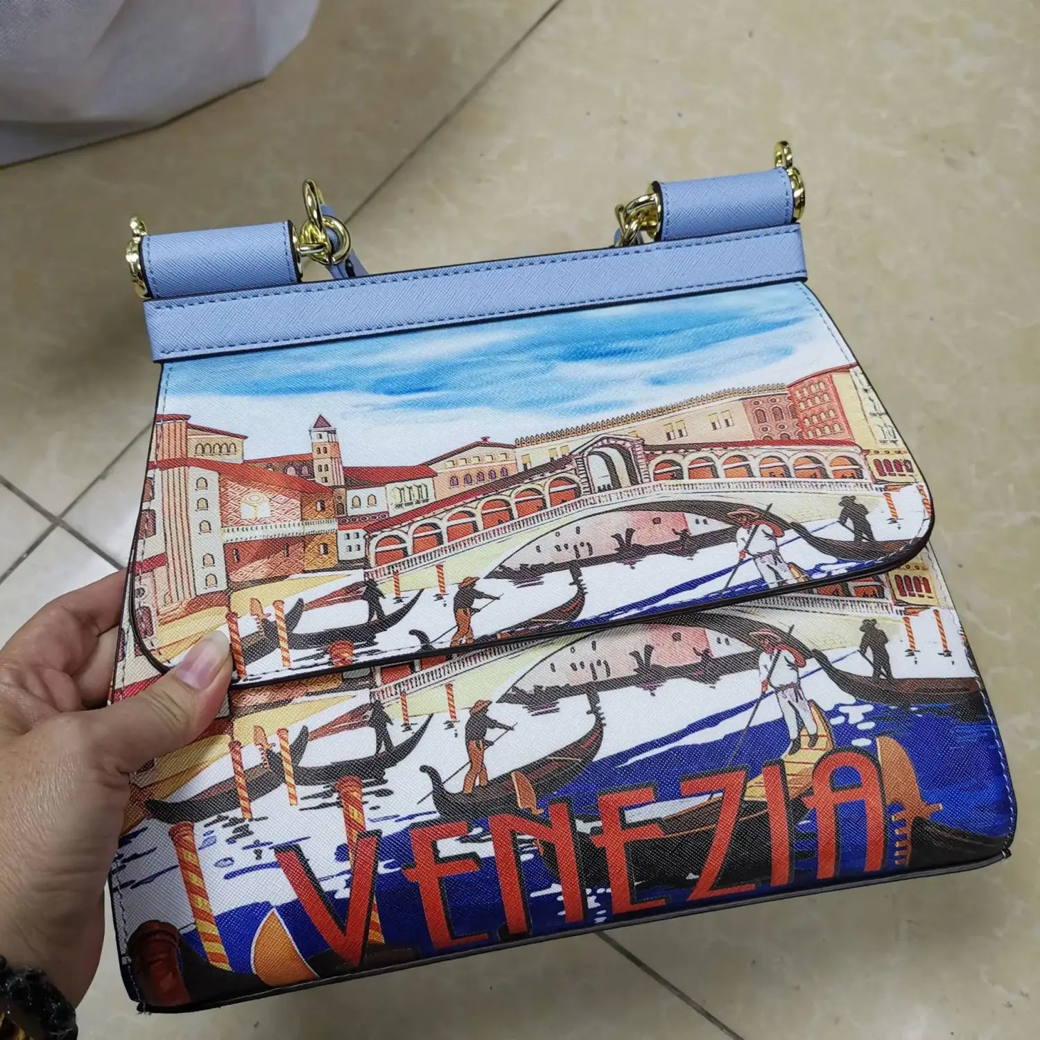 Sac bandoulière en cuir vintage bleu clair, art raffiné, avec un design illustré vibrant de Venise, des gondoles et des bâtiments sous un ciel accentué par une quincaillerie ton or.