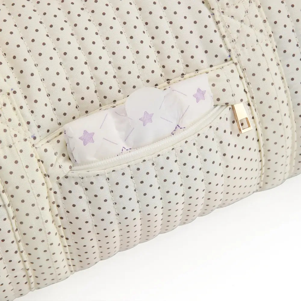 Eine cremefarbene gepolsterte Kinderwagen-Organizer-Tasche mit schwarzen Pünktchen und einem goldfarbenen Zipper-Anhänger mit Teddybär-Motiv.