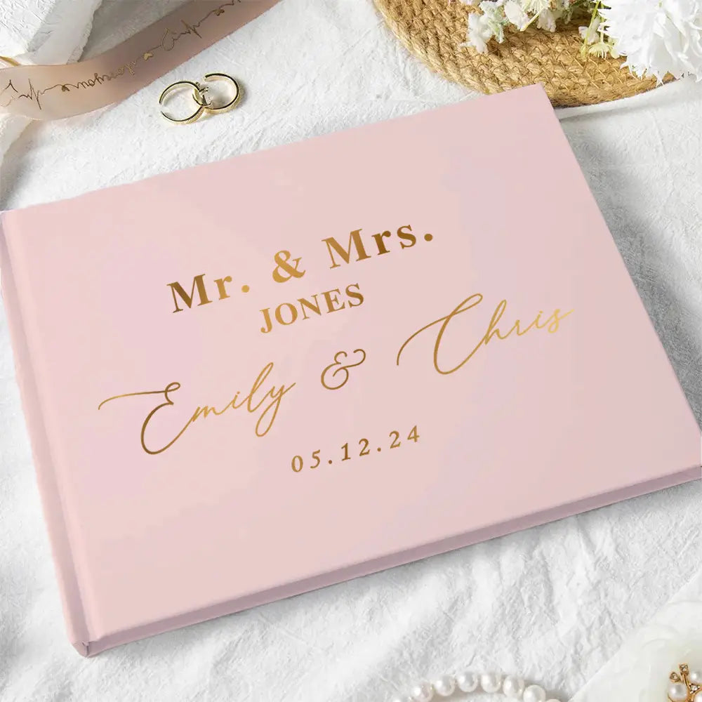 Personalized pink wedding guest book with gold foil lettering and elegant script design accompanied by a pen for signing.