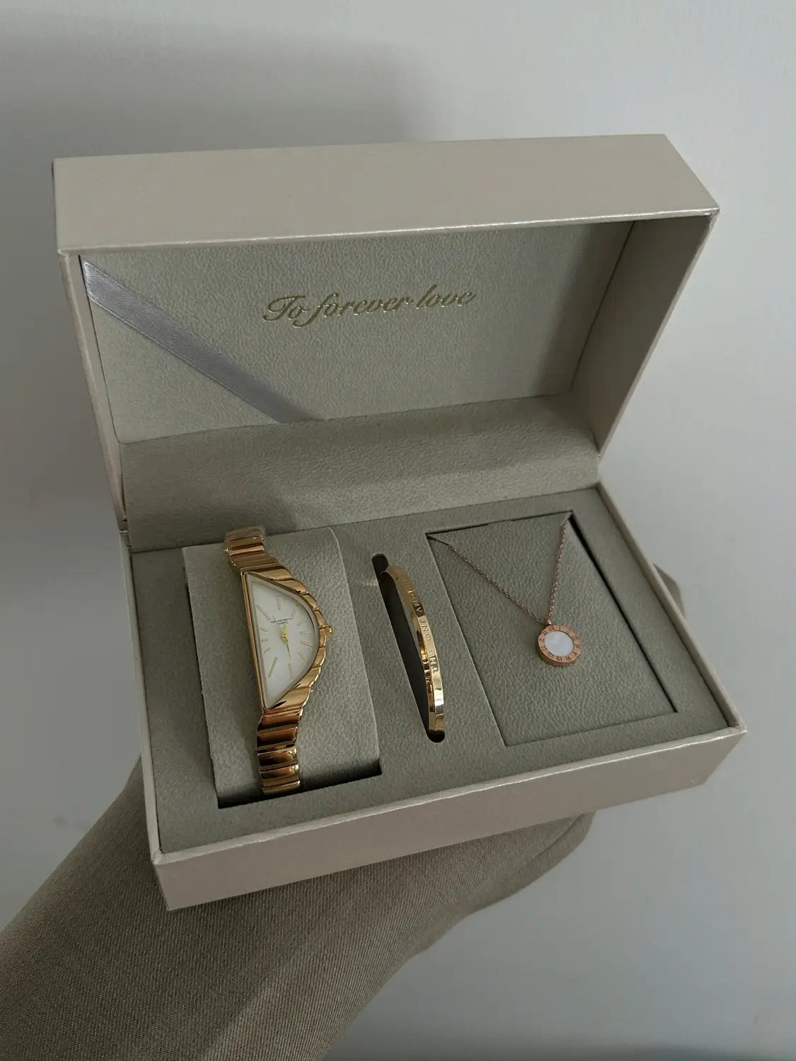 gold-toned geometric triangle quartz watch with a curved rectangular face and stainless steel linked bracelet displayed in a beige gift box alongside matching jewelry