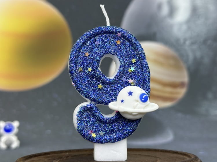 Una vela azul con brillo con el número nueve para cumpleaños, con una figura de astronauta blanca adherida a su curva inferior, diseñada como adorno para pastel de fiesta planetaria.